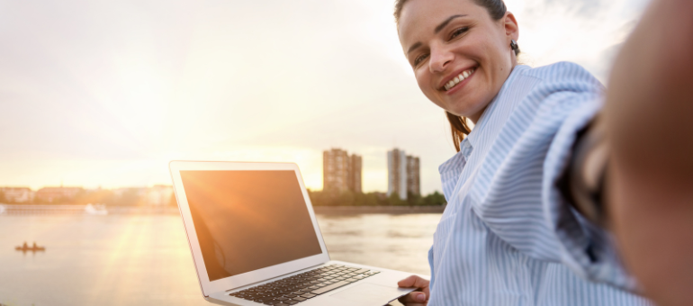 Eine Frau sitzt am Ufer, hat ihren Laptop aufgeklappt auf ihrem Schoß und macht ein Selfie im Sonnenuntergang