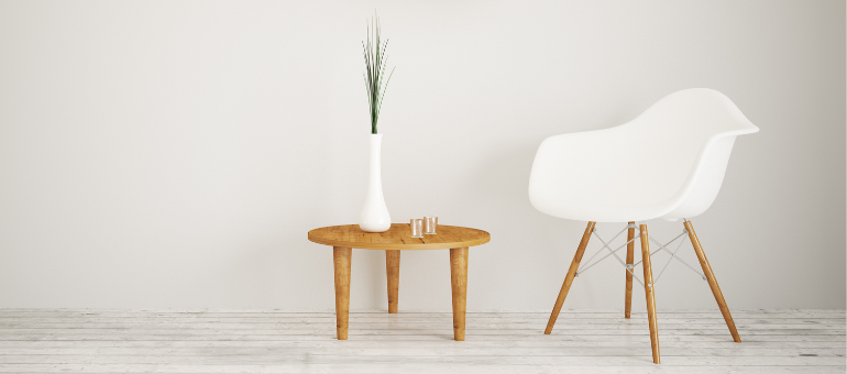 Ein weißer Stuhl steht neben einem kleinen Holztisch. Auf diesem steht eine weiße, dünne Vase mit grünen Trockenblumen