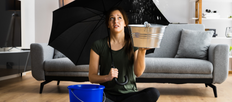 Eine Frau sitzt im Schneidersitz auf dem Boden in ihrem Wohnzimmer, mit einem Regenschirm in der Hand und einem Eimer neben sich stehen. Sie sieht traurig aus und es tropft über ihr von der Decke..