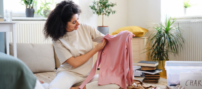 Eine Frau  sitzt in ihrem Wohnzimmer auf dem Boden und hält lächelnd einen rosa Pullover nach oben. 