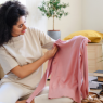 Eine Frau  sitzt in ihrem Wohnzimmer auf dem Boden und hält lächelnd einen rosa Pullover nach oben. 